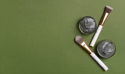 Bowls of activated carbon facial mask and makeup brushes on green background