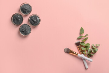 Bowls of activated carbon facial mask, powder, makeup brushes and eucalyptus branch on pink...
