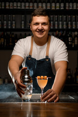 Young smiling barman professionally holds transparent shaker with ice cubes and glass of cold alcoholic cocktail