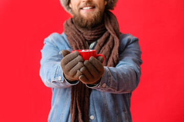 Young bearded man in warm gloves with cup of cocoa on red background