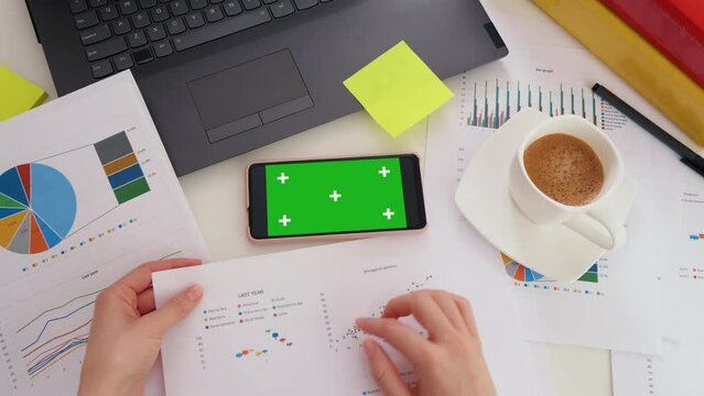 Top view of smartphone with green screen on the office desk. Application for business. Laptop, worksheet finance, graph accounts on office table workspace. Chroma key. Analysis of financial report