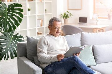 Senior man in eyeglasses reading magazine on sofa at home