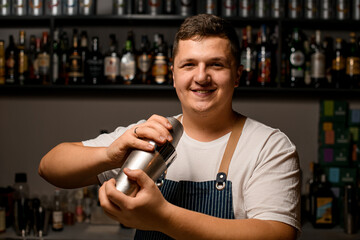 Young smiling bartender shaking a cocktail shaker in bar interior