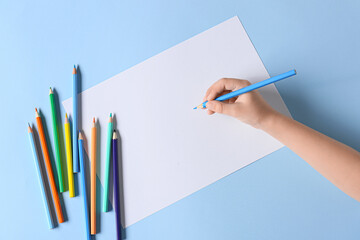 Child's hand with blank sheet of paper and different pencils on color background