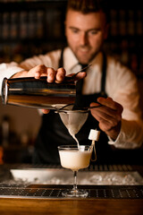 man bartender carefully filters bright alcoholic cocktail from steel shaker cup into glass through sieve. Close-up view.