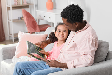 Happy African-American man and his little daughter reading book at home
