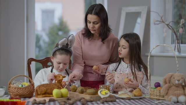Concentrated Caucasian mother and daughters coloring eggs on Easter at home indoors. Front view portrait of beautiful woman and charming girls preparing traditional food on spring holiday
