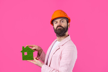 Portrait of young construction engineer wear orange hard hat, in a pink jacket standing on red studio background. A man holds a green eco house.