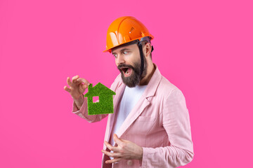 Portrait of young construction engineer wear orange hard hat, in a pink jacket standing on red studio background. A man holds a green eco house.