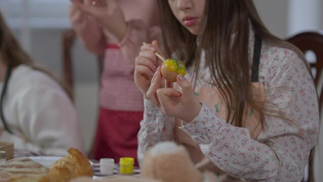 Unrecognizable teenage girl coloring Easter egg with paint as blurred sister and mother preparing dinner at background. Caucasian teenager painting on traditional holiday food at home indoors