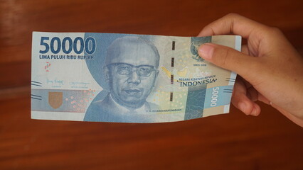 The Indonesian currency, the 50,000 rupiah note, is held in a woman's hand