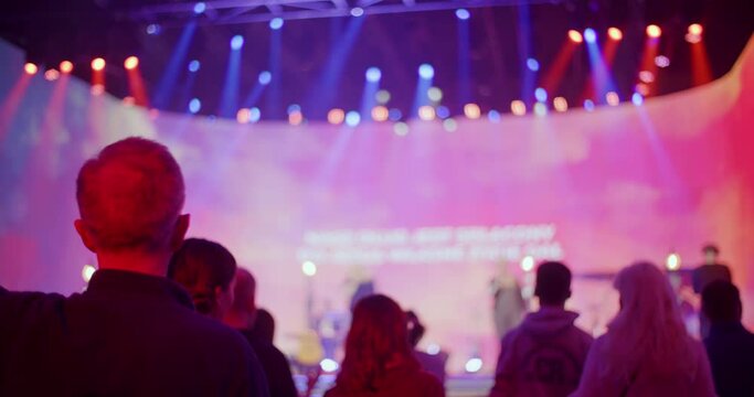 Christians Worship and Praise God on Music Concert, Youth Festival or Conference. Unrecognizable People Sing Songs with Raised Arms on Live Stage Event with Lights. 4K backside view handheld shot