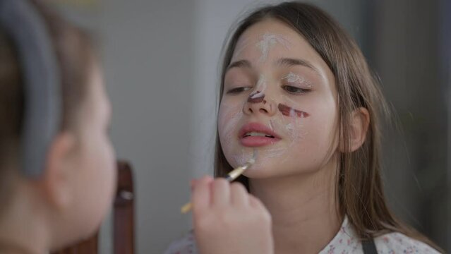 Close-up portrait of beautiful teenage girl indoors with little sister painting face with food coloring. Caucasian positive teenager having fun with sibling at home on Easter Sunday. Joy and family