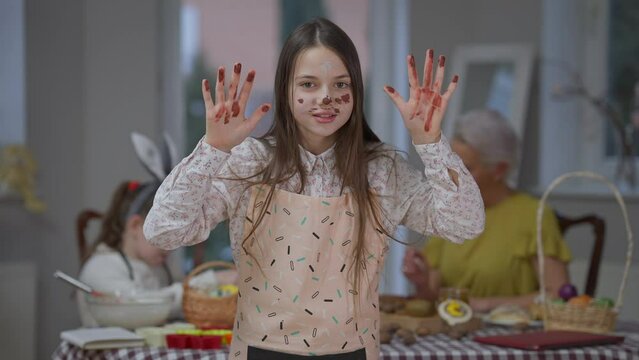 Portrait of positive teenage girl with colored hands and face looking at camera gesturing. Medium shot of joyful Caucasian teen posing at home with blurred family sitting at Easter dinner table