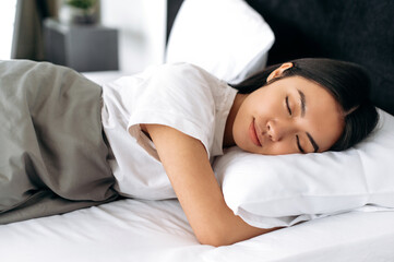 Sweet dreams. Pretty chinese woman in white t-shirt under the blanket in bed in the morning. Side view of a sleeping asian girl in a cozy bed in apartment, pleasant dreams, healthy sleep