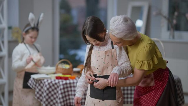 Smiling beautiful grandmother tying apron on teenage granddaughter with little girl cooking at background. Happy Caucasian family preparing Easter dinner at home indoors