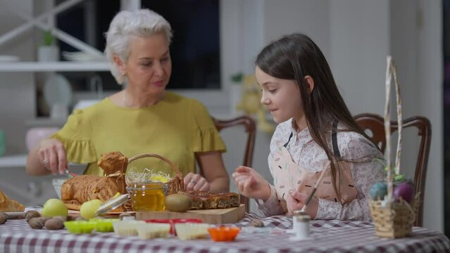 Carefree teenage girl eating delicious honey with finger as blurred woman scolding at background. Portrait of Caucasian granddaughter enjoying tasty dessert as grandmother cooking Easter dinner