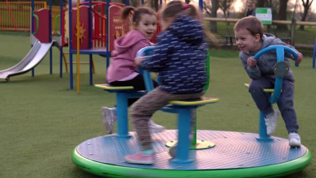 Three little Happy Children having fun spinning on merry-go-round. Smiling Siblings friends playing outside. emotional kids rides on carousel at playground nursery in park.Happy family, childhood