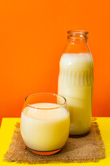 Glass bottle and big glass with milk on a square of rustic fabric on a yellow table with red background. Copy space.