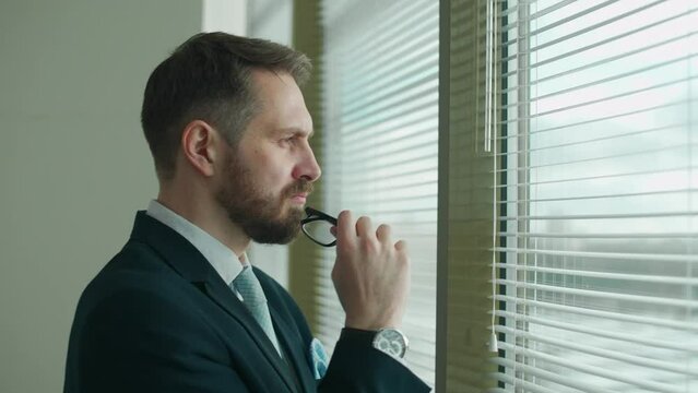 Focused businessman in a suit stands at the panoramic window in the office and reflects, the thought process, natural lighting.