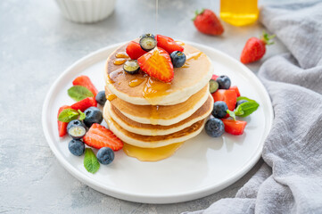 A stack of delicious fluffy pancakes with berries and honey on a white plate on a gray concrete background. Copy space.