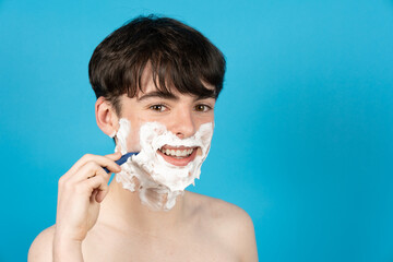 Happy teenager boy shaving face with foam and razor.