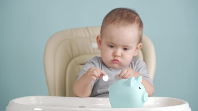 Baby 12-17 months throws a Russian ruble coin into a blue piggy bank sitting on a chair, the concept of maternity capital