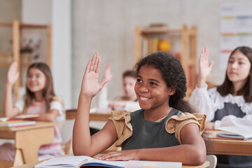 Portrait of smiling African-American schoolgirl raising hand while sitting at desk in classroom, copy space