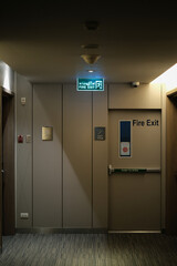 Emergency exit sign at the corridor in building. Green fire exit sign hanging on ceiling on dark corridor in building near fire emergency exit door. Green emergency exit sign.