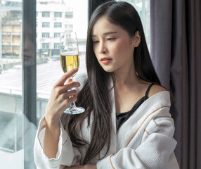 Beautiful woman in bathrobe with glass of champagne in bedroom. Young woman drinking champagne standing by the window in hotel room. Young Asian woman drinking champagne.