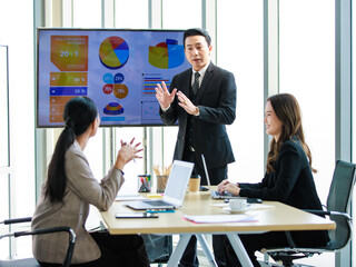 Asian professional successful businessman in formal suit standing showing presenting explaining report investment graph chart data from computer monitor in meeting room to businesswoman colleagues