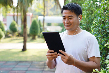 Fototapeta premium Asian man listens and watch video clips on smart tablet at the park. Concept : Give time for yourself to relax with wireless technology. Internet of things. Social media. 