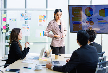 Asian professional successful businesswoman in formal suit standing showing presenting explaining report investment graph chart data from computer monitor in meeting room to business team colleagues