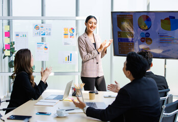 Asian professional successful businesswoman in formal suit standing showing presenting explaining report investment graph chart data from computer monitor in meeting room to business team colleagues