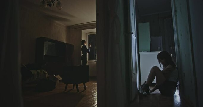 Two young women relaxing in their apartment at night. Two young women relaxing in their apartment at night in different rooms, one smoking and one drinking