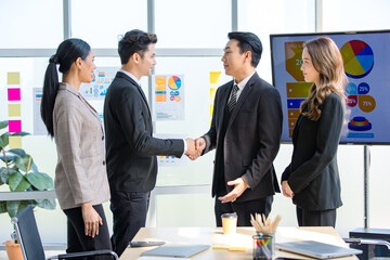 Asian young happy professional successful businessman and businesswoman partnership standing shaking hands greeting together while having job agreement achievement deal done in company meeting room