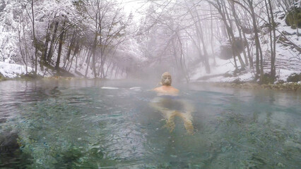 A man in a hat swimming in a natural thermic spring in Maibachl, Austria during snowfall, winter....