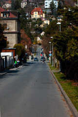 Urban perspective, a street that goes down and up. View of a street in a residential area, with many houses  gardens and trees. The road is straight and goes down and then up towards a house red roof