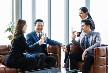 Group of Asian happy cheerful professional successful businessmen and businesswomen in formal suit sitting on leather sofa smiling holding tall champagne glass toasting celebrating cheers together
