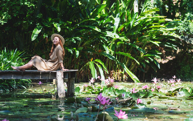 Portrait image of a beautiful young asian woman sitting on wooden bridge in a pond with pink lotus flowers