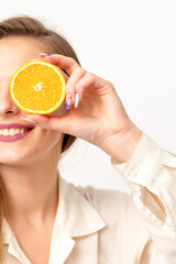 Portrait of a cheerful caucasian young woman covering eye with an orange slice wearing a white shirt over a white background