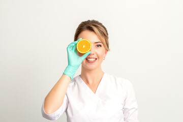Portrait of young caucasian smiling female beautician covering eye with an orange slice wearing gloves over a white background