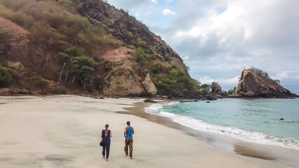 A couple walking on an idyllic Koka Beach. Hidden gem of Flores, Indonesia. Couple is enjoying their romantic escape. Waves gently washing the shore. There are hills in the back. Happiness and love