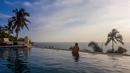 A young man leaning at the edge of an infinity pool and looking at the sun setting into the sea. There is a big palm tree in front. Calmness and serenity. Luxury hotel in Lombok, Indonesia.