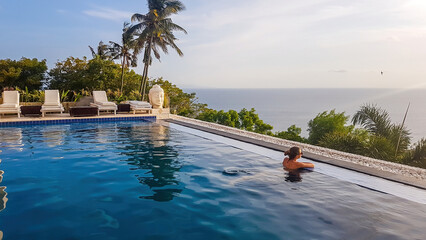 A young woman leaning at the edge of an infinity pool and looking at the sun setting into the sea....