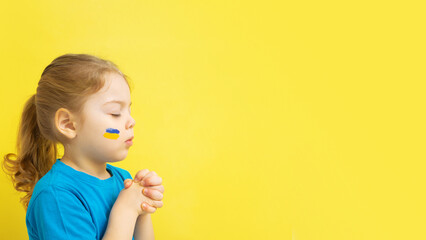 girl holding folded hands in prayer with yellow and blue colors of the Ukrainian flag on his cheek
