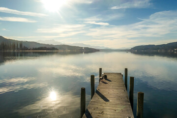 A wooden pier going into the lake, with Alps in the back. The calm surface of the lake is reflecting the mountains, sunbeams and clouds. Clear and sunny day. Few ducks standing at the promenade