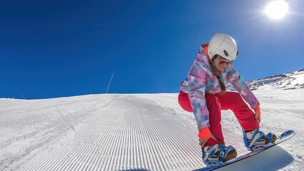A snowboarding girl in pink trousers and helmet going down a perfectly groomed slope. She is...