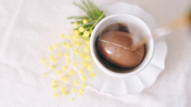 Top view of hot coffee is poured on a brown painted Easter egg in a white cup breaking it on white table background.