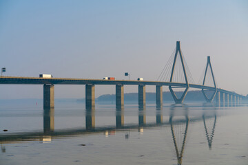 Long highway bridge spanning over a bay, with traffic driving on it. Calm water and morning haze.